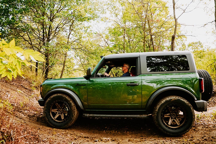 A green Ford Bronco driving on a dirt path in a forest