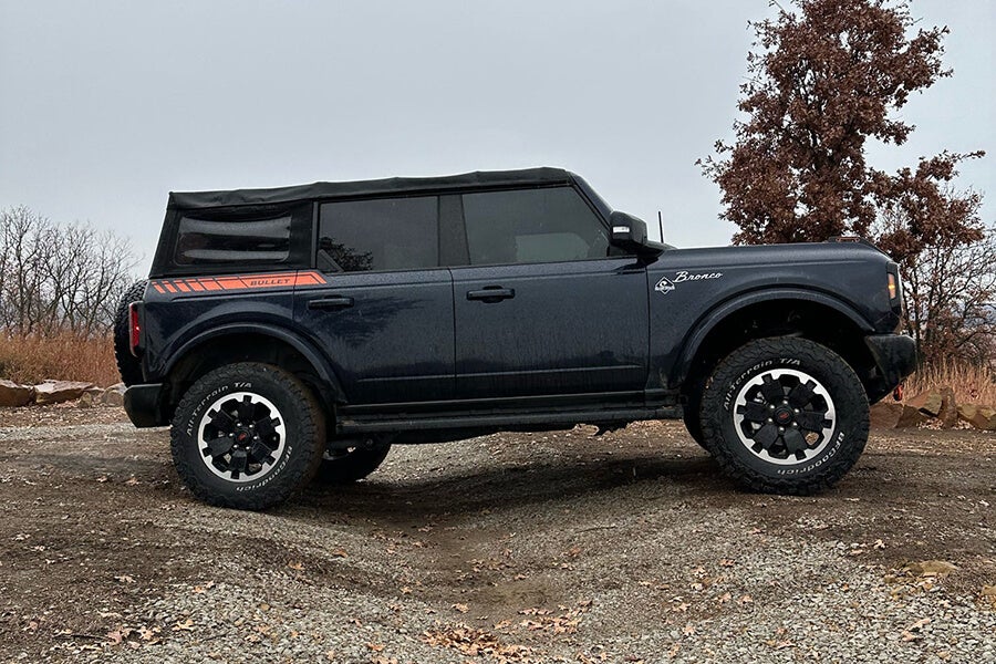 A dark gray Ford Bronco with a black soft top parked on a dirt road.