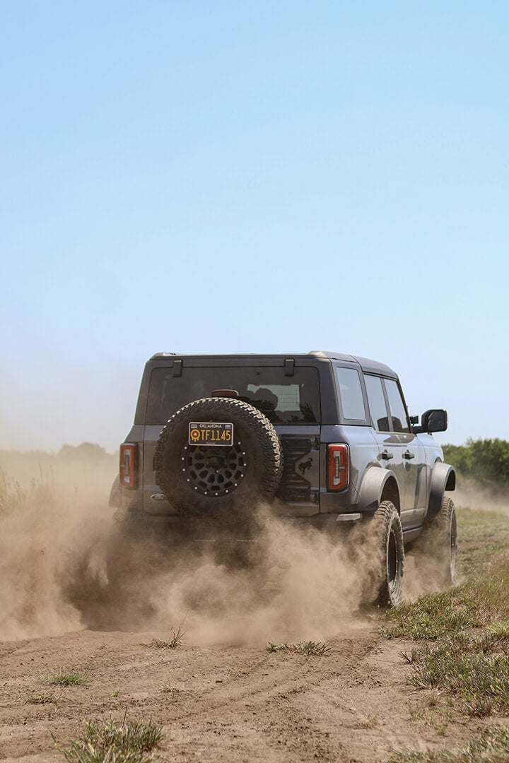 A rear view of a Ford Bronco with a tilted tire, kicking up dust.