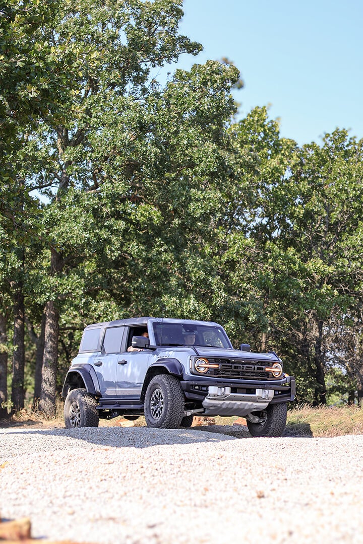 A side view of a blue Ford Bronco driving on a dirt road.