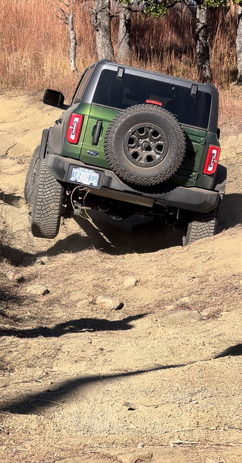 A close-up of a Ford Bronco's rear tire and suspension on uneven ground