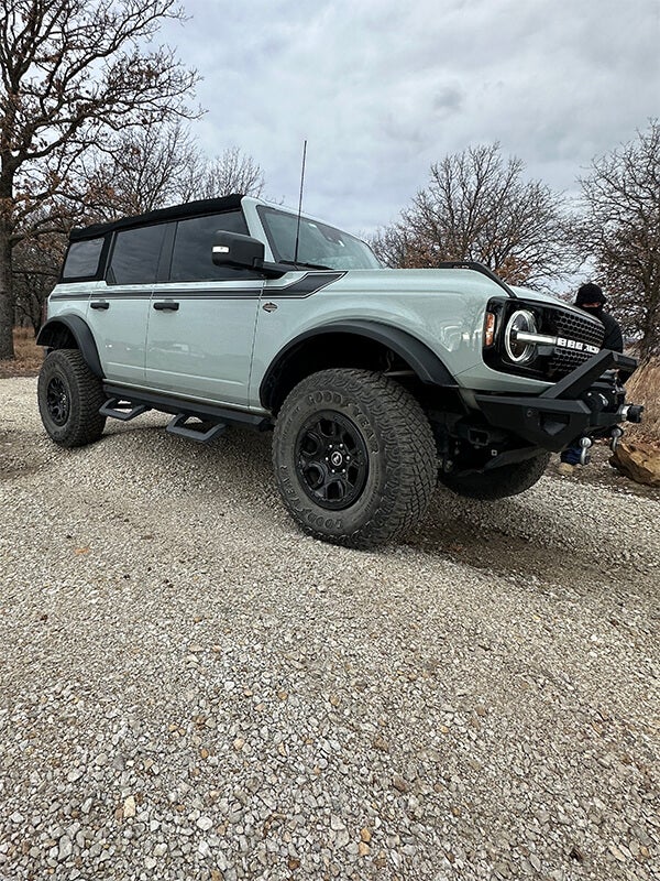 A two-door white Ford Bronco with black stripes parked on gravel