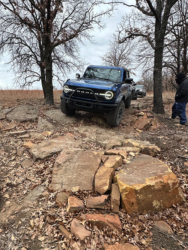 A front view of a blue Ford Bronco driving over large rocks