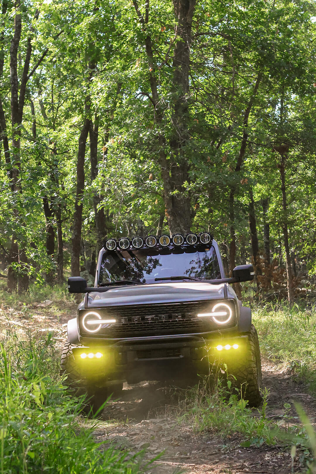 A front view of a dark Ford Bronco driving on a dirt trail.
